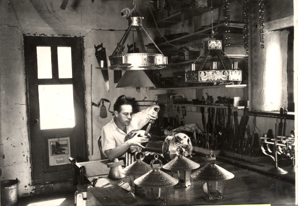 Robert Woodman, in his Canyon Road workshop surrounded
by numerous completed items and works in progress, solders a
large cylindrical fixture designed to utilize tubular fluorescent
light bulbs, circa 1940-1950. Private collection.